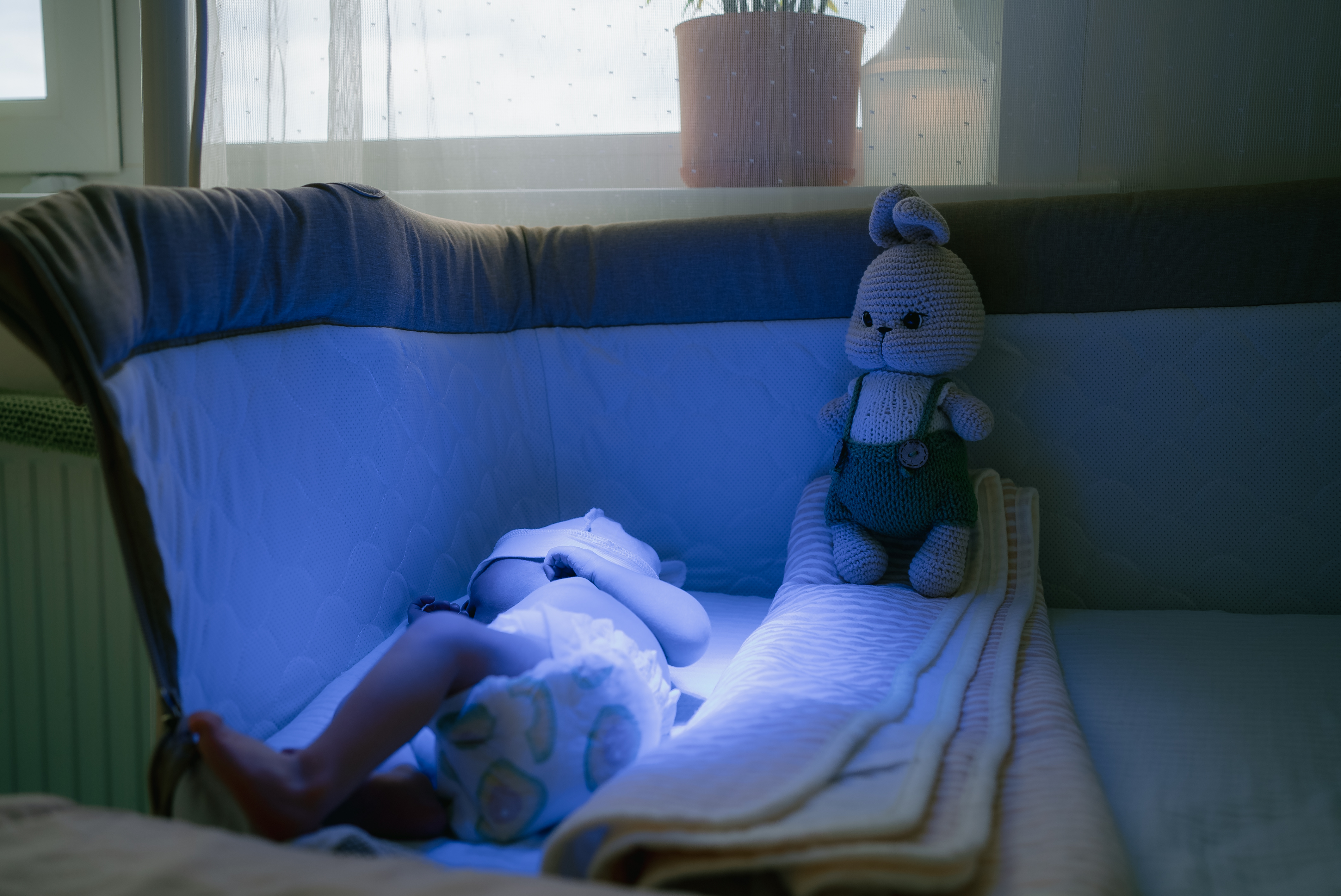 Recém-nascido descansando em um berço médico sob luz azul de fototerapia, com proteção ocular e um ursinho ao lado